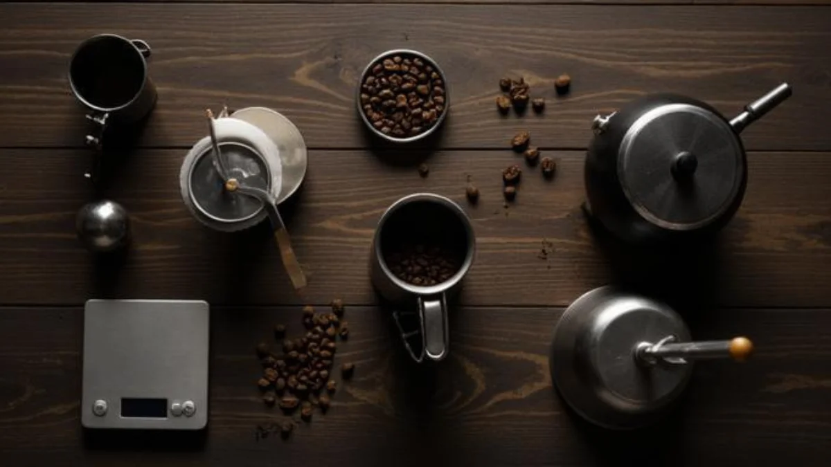 A flat lay of beginner coffee equipment including a V60 dripper, French press, manual grinder, scale and whole coffee beans on a dark wooden surface