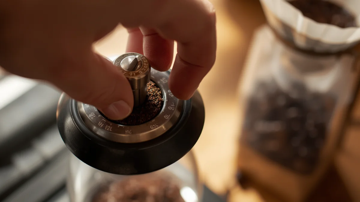 Hand adjusting grind size dial on a manual burr grinder with coffee grounds and pour over equipment in the background